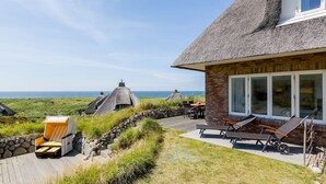 Una casa costera con techo de paja, pared de piedra y un patio con sillones de descanso.