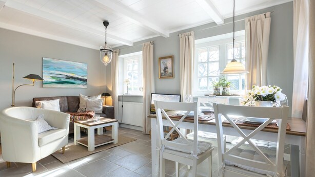 A well-lit living room with a white sofa, a coffee table, and a dining area with white chairs and a table set for two. - Paulas Hüs - Fiete - Die Fiete, ca. 50 m² ist Besonders Liebevoll für bis zu 3 Personen (Sylt-Ost)