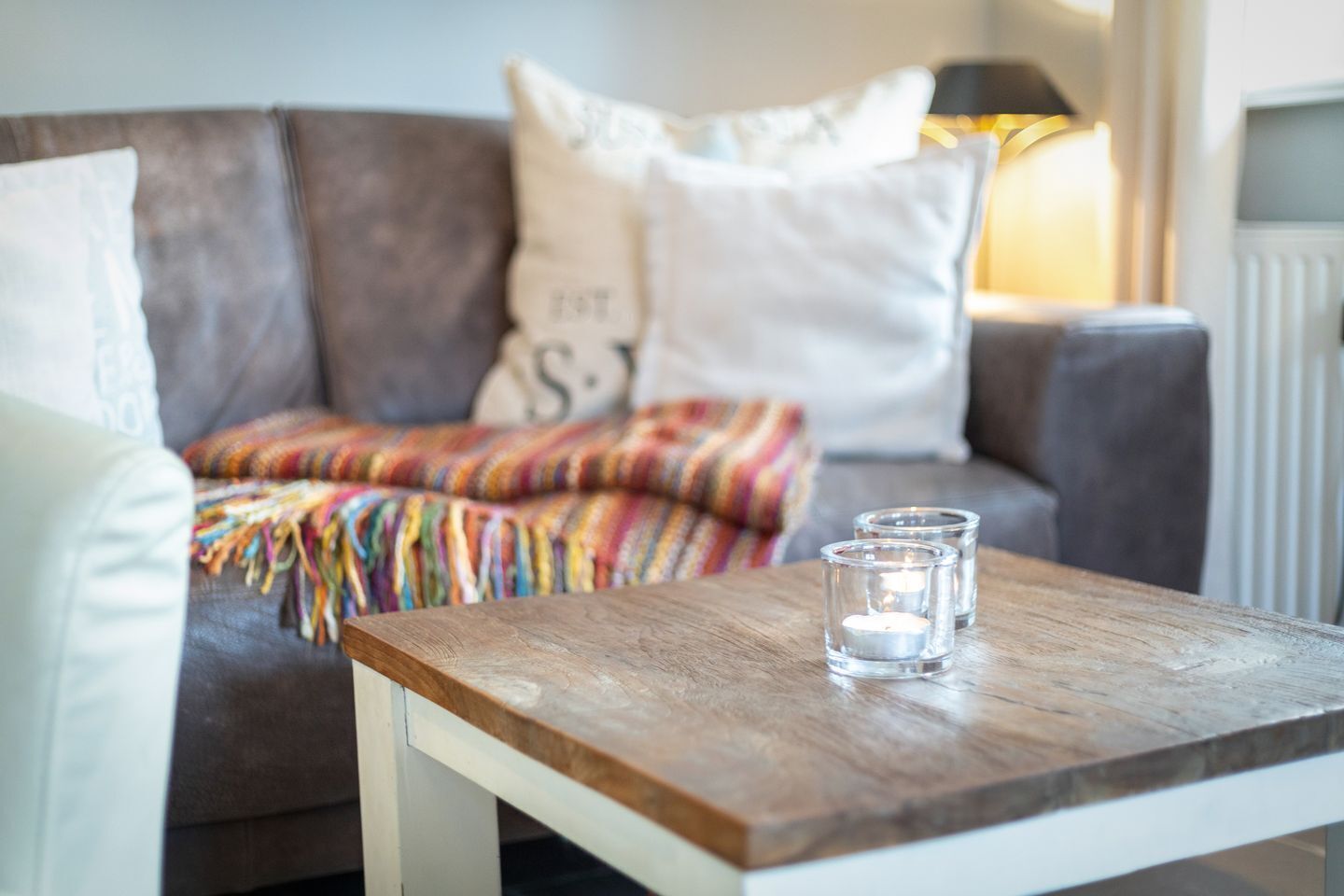 Ein gemütliches Wohnzimmer mit einer braunen Couch, weißen Kissen, einer farbenfrohen Decke und einem Holztisch mit zwei Glaswindlichtern.
