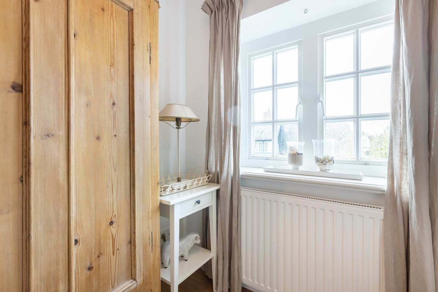 A room with a wooden wardrobe, a white bedside table with a lamp, a white radiator, and a window with curtains.