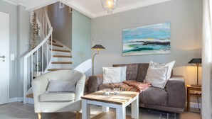 A living room with a staircase, a sofa set, a coffee table, and a painting on the wall. - Paulas Hüs - Fiete - Die Fiete, ca. 50 m² ist Besonders Liebevoll für bis zu 3 Personen (Sylt-Ost)