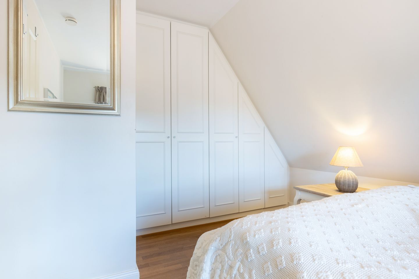 Un dormitorio con techo inclinado, armario blanco, lámpara de noche y cama con cubrecama blanco texturizado.
