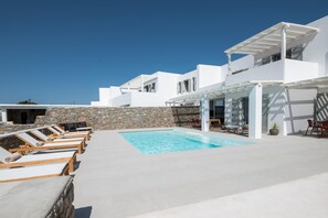 A modern outdoor pool area with lounge chairs, a stone wall, and a white building with a pergola. - Petra Mare in Mykonos (Mykonos)