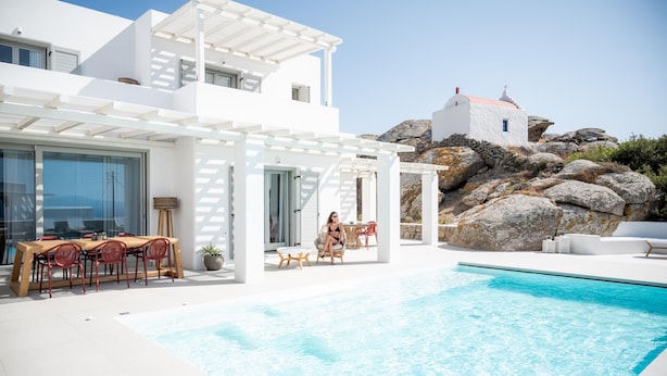 A modern white villa with a swimming pool, outdoor seating, and a view of a rocky landscape and a church. - Petra Mare in Mykonos (Mykonos)