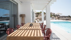 A wooden table with chairs in a covered outdoor area with a pool in the background. - Petra Mare in Mykonos (Mykonos)