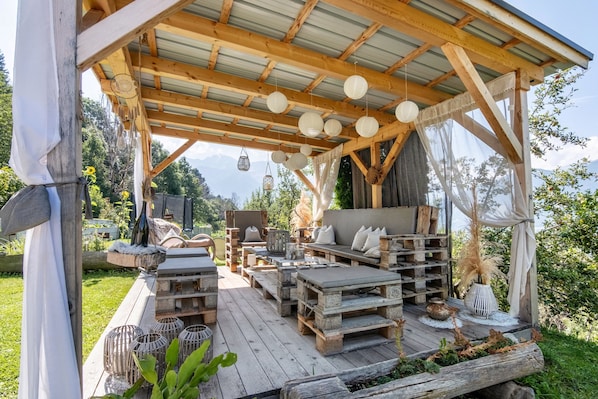 Une terrasse extérieure en bois, meublée de façon rustique, avec un espace salon abrité et une vue sur les montagnes.