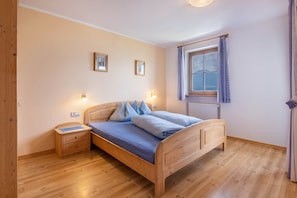 Un lit en bois avec un matelas bleu, une table de chevet en bois, deux lampes fixées au mur et une fenêtre avec des rideaux.