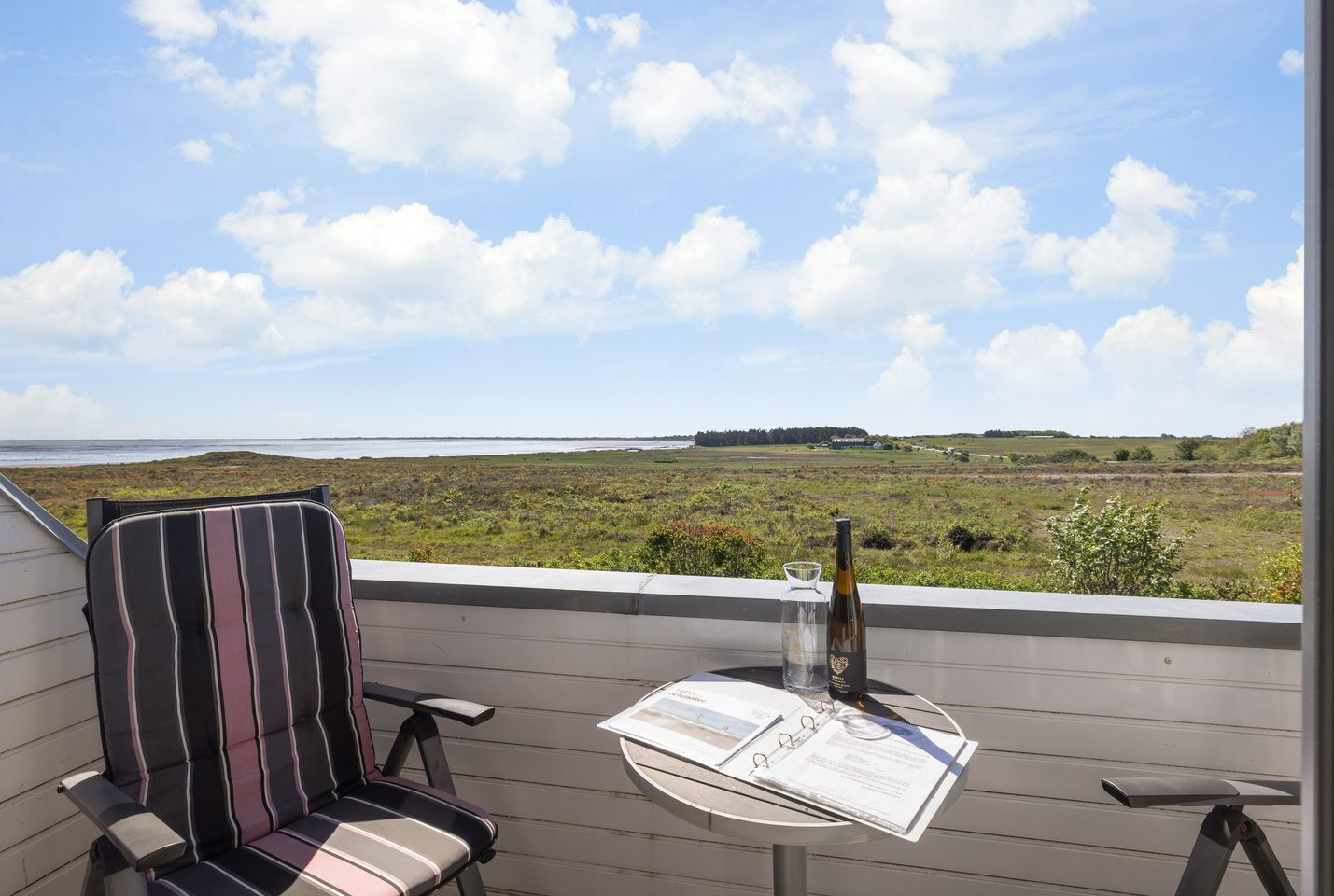 A balcony with a striped chair, a small table with a bottle and a glass, and a view of a grassy landscape and water.