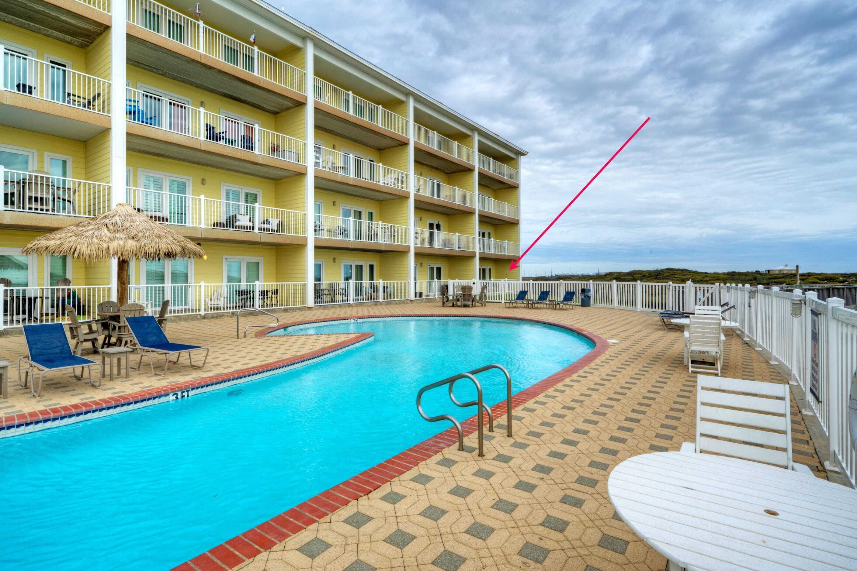 A swimming pool with lounge chairs, a thatched umbrella, and a multi-story building with balconies.