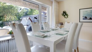 Un coin repas avec une table dressée pour six personnes, des chaises blanches, une vue sur l’extérieur et un tableau encadré au mur.