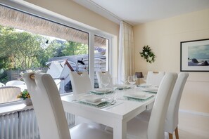 Un coin repas avec une table dressée pour six personnes, des chaises blanches, une vue sur l’extérieur et un tableau encadré au mur.