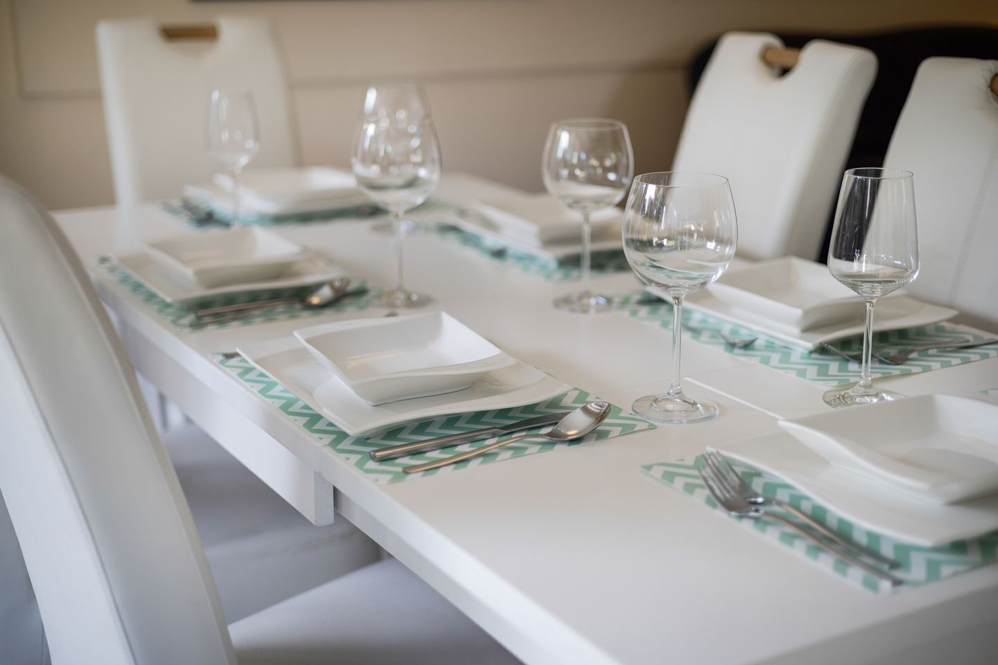 Una mesa dispuesta con platos blancos, cubiertos y copas de cristal, sobre un mantel blanco y posavasos con motivos verdes.