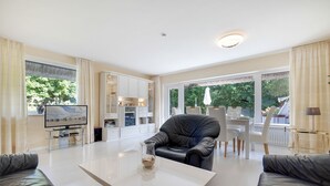 A modern living room with a flat-screen TV, a glass coffee table, a leather armchair, and a dining area with white chairs.