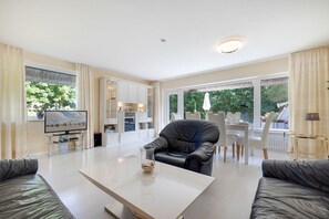 A modern living room with a flat-screen TV, a glass coffee table, a leather armchair, and a dining area with white chairs.