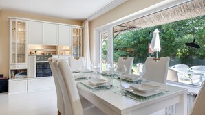 A dining area with a table set for four, white chairs, and a view of a garden through large windows.