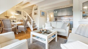 A spacious living room with a white sofa, a coffee table, and a dining area with wicker chairs. A staircase leads to an upper level. - Elbe, Wohnung Nr. 8 - Haus Elbe - Liebevoll Ausgestattete Obergeschoss-wohnung Unter Reet (Sylt-Ost)