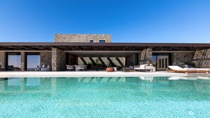 A modern outdoor pool area with a stone facade building, outdoor seating, and a clear blue pool. - Villa Sky II in Mykonos (Mykonos)