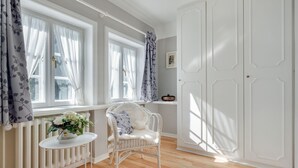 A room with a white radiator, a wicker chair, a small round table with a plant, and a white wardrobe.