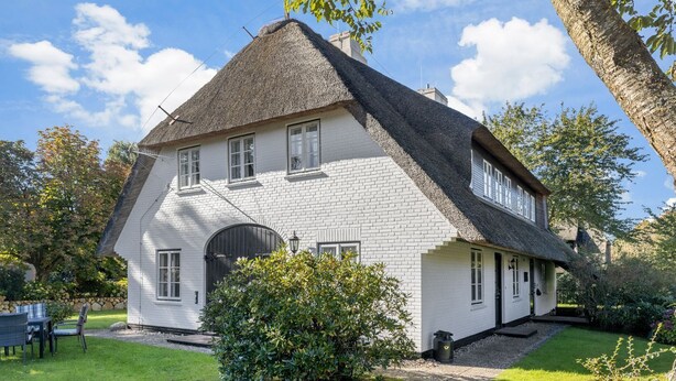 A traditional house with a steeply pitched roof, white brick walls, and a small garden with a lawn and a bush.