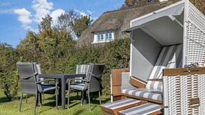 A cozy outdoor seating area with wicker furniture and a covered sleeping area, set against a thatched-roof house and lush greenery.