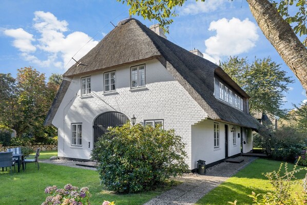 Ein traditionelles weißes Haus mit einem steil geneigten Strohdach, einem markanten Rundbogenportal und einem gepflegten Garten mit verschiedenen Pflanzen und Blumen.