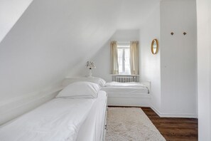 Ein kleines, weißes Schlafzimmer im Mansardenstil mit zwei Einzelbetten, einem weißen Heizkörper, einem Fenster mit Vorhängen und einem Spiegel an der Wand.