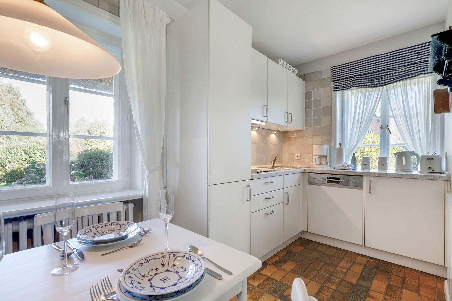 Una cocina moderna con armarios blancos, una mesa de comedor dispuesta con platos y cubiertos, y una ventana con cortinas.