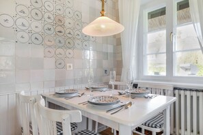 Comedor con una mesa blanca dispuesta para cuatro personas, rodeada de sillas blancas con cojines a cuadros. Se observa una pared con platos circulares de diferentes diseños detrás de la mesa.
