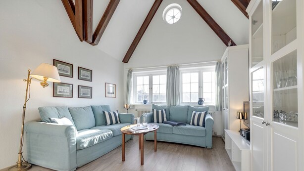 A living room with a light blue sofa, a wooden coffee table, and a floor lamp. There are framed pictures on the wall and a window with curtains. - Giebel, Alte Mühle - Alte Mühle Giebel - in Erster Reihe zum Wattenmeer (Sylt)