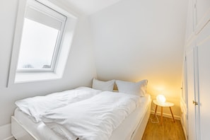 Una cama individual con ropa blanca en una habitación pequeña de paredes blancas, con tragaluz y mesita de noche con lámpara.