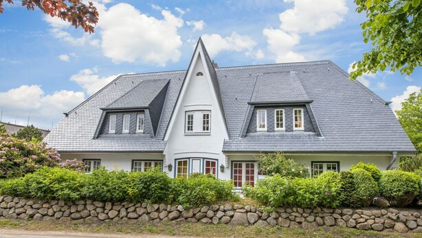 A two-story house with a steeply pitched roof, a stone wall in front, and a clear blue sky. - Süder Ech - Süder Ech - ein Gepflegtes Haus, Zentral Gelegen (Sylt)