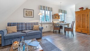 A cozy living room with a blue sofa, a dining table with chairs, a wooden cabinet, and a window with striped curtains. - Süder Ech - Süder Ech - ein Gepflegtes Haus, Zentral Gelegen (Sylt)