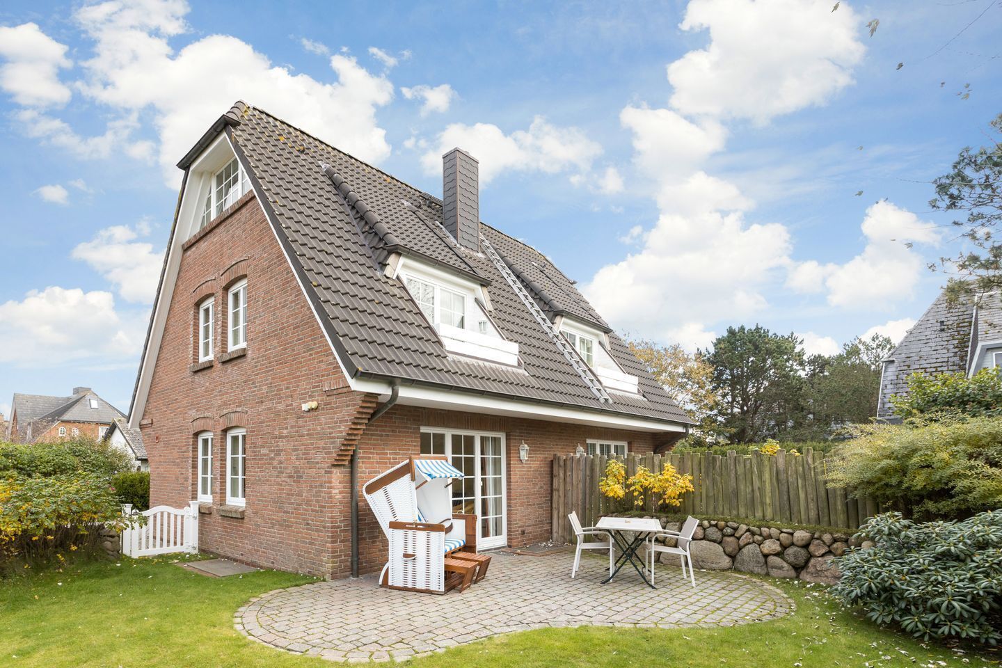 A two-story brick house with a sloped roof, a small patio with a table and chairs, and a white fence surrounding the property.