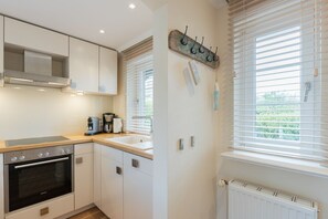 A modern kitchen with white cabinets, a stainless steel oven, a coffee maker, and a window with blinds. - Barig Hüs EG - Barig Hüs EG - Objekt in Einer Ruhig Gelegenen Straße in der Nähe vom Wattenmeer (Sylt-Ost)