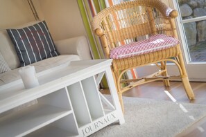 A wicker chair with a cushion, a white side table, and a sofa with striped pillows. - Barig Hüs EG - Barig Hüs EG - Objekt in Einer Ruhig Gelegenen Straße in der Nähe vom Wattenmeer (Sylt-Ost)
