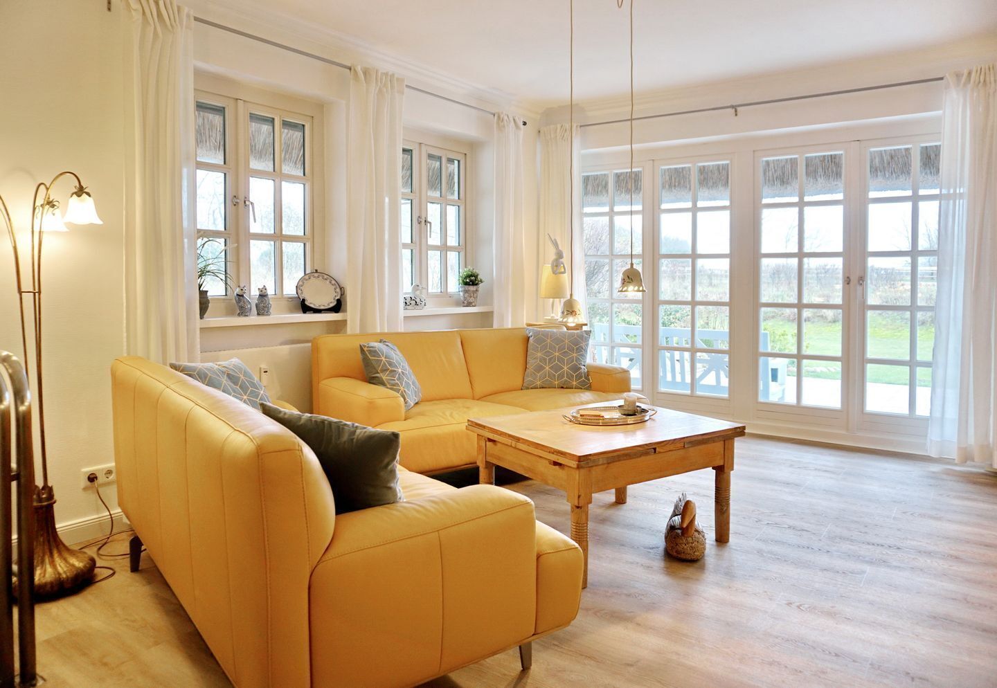 Ein geräumiges Wohnzimmer mit einem gelben Sofa, einem Holztisch und großen Fenstern mit Blick ins Freie.