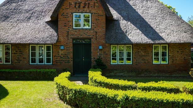 A traditional brick house with a steeply pitched thatched roof, a central entrance with double doors, and a small garden with neatly trimmed hedges.