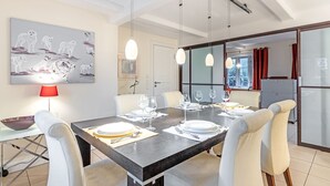 A dining area with a large table set for four, white chairs, pendant lights, and a red table lamp.