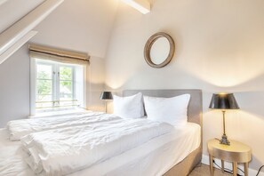 Une chambre avec un grand lit, deux tables de chevet avec des lampes, une fenêtre avec des stores et un grand miroir rond accroché au mur.