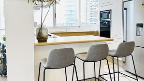 A modern kitchen with a large window, a built-in oven, and a refrigerator. There are three bar stools and a potted plant on the counter. - Beach View 2 Bedrooms Apartment  (Surfers Paradise)