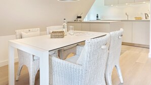 Un'area pranzo moderna con tavolo bianco e sedie in vimini, una lampada a sospensione e una cucina sullo sfondo.