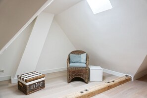 Une petite pièce aux murs inclinés, avec un fauteuil en osier, une armoire blanche et une commode en osier.