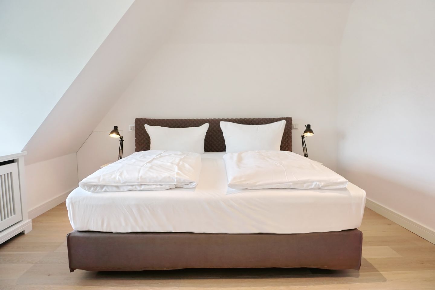 A neatly made bed in a room with a sloped ceiling, two bedside lamps, and a radiator.