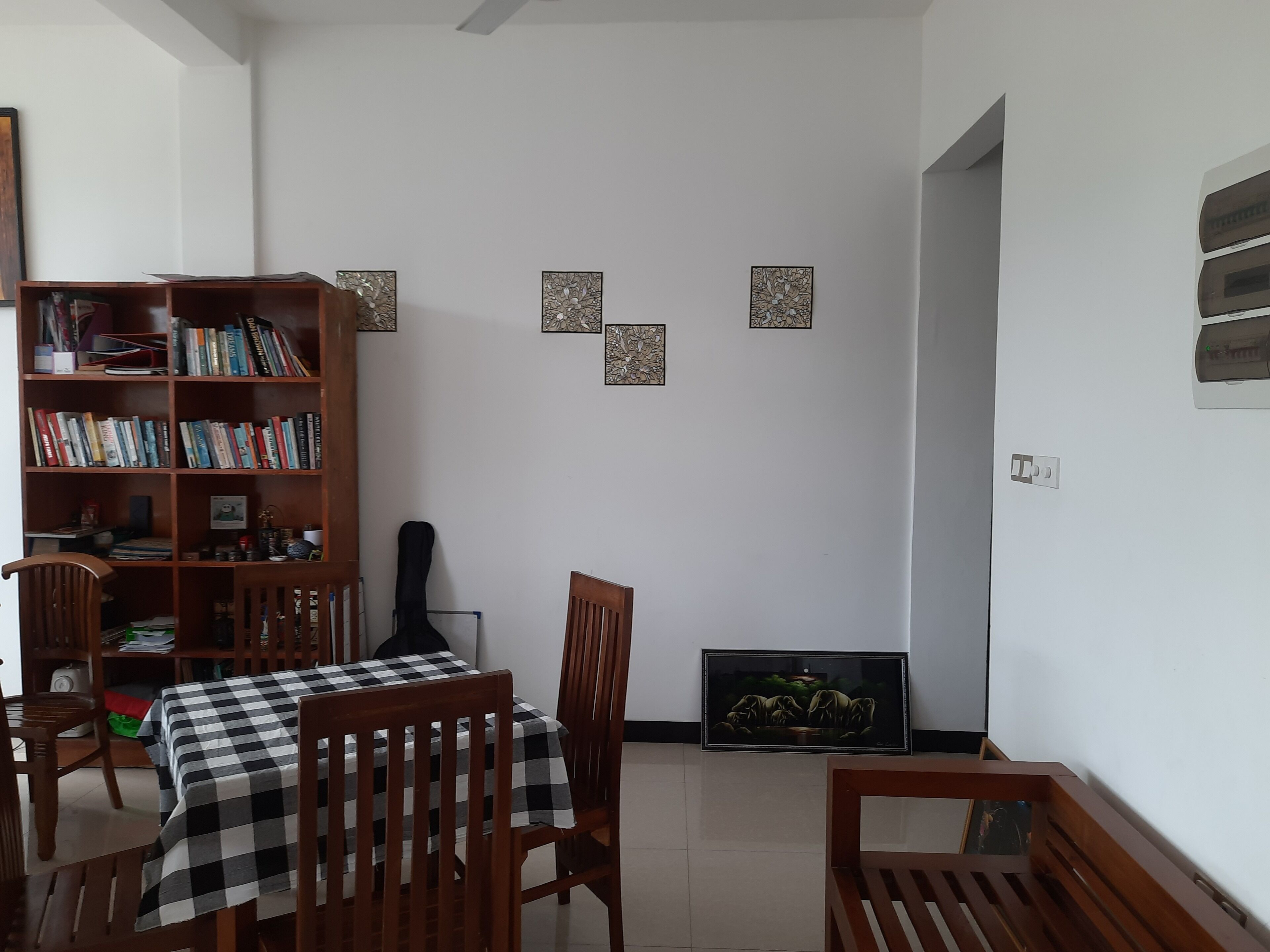 A dining area with a checkered tablecloth, wooden chairs, a bookshelf, and decorative wall art.