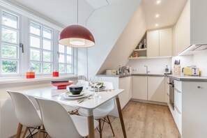 A modern kitchen with white cabinetry, a dining area with white chairs, and a large window with natural light. - Auszeit im Takerhof - Auszeit im Takerhof - Ferienwohnung in Wunderschöner Südlage in Keitum (Sylt-Ost)
