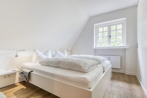 A white, modern bedroom with a sloped ceiling, a bed with white bedding, a bedside table, a lamp, and a window with a view of greenery. - Auszeit im Takerhof - Auszeit im Takerhof - Ferienwohnung in Wunderschöner Südlage in Keitum (Sylt-Ost)