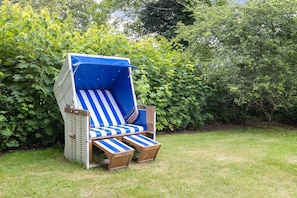A blue and white striped outdoor chair with a metal frame and wooden armrests, placed on a grassy lawn with dense green foliage in the background. - Auszeit im Takerhof - Auszeit im Takerhof - Ferienwohnung in Wunderschöner Südlage in Keitum (Sylt-Ost)