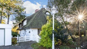 Une maison traditionnelle à toit de chaume, avec une façade blanche, une clôture en bois peinte en blanc et un chemin pavé menant à l’entrée.