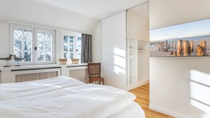 Une chambre moderne avec un grand lit, une fenêtre donnant sur des arbres, un fauteuil et une armoire ornée d’un tableau représentant une plage.