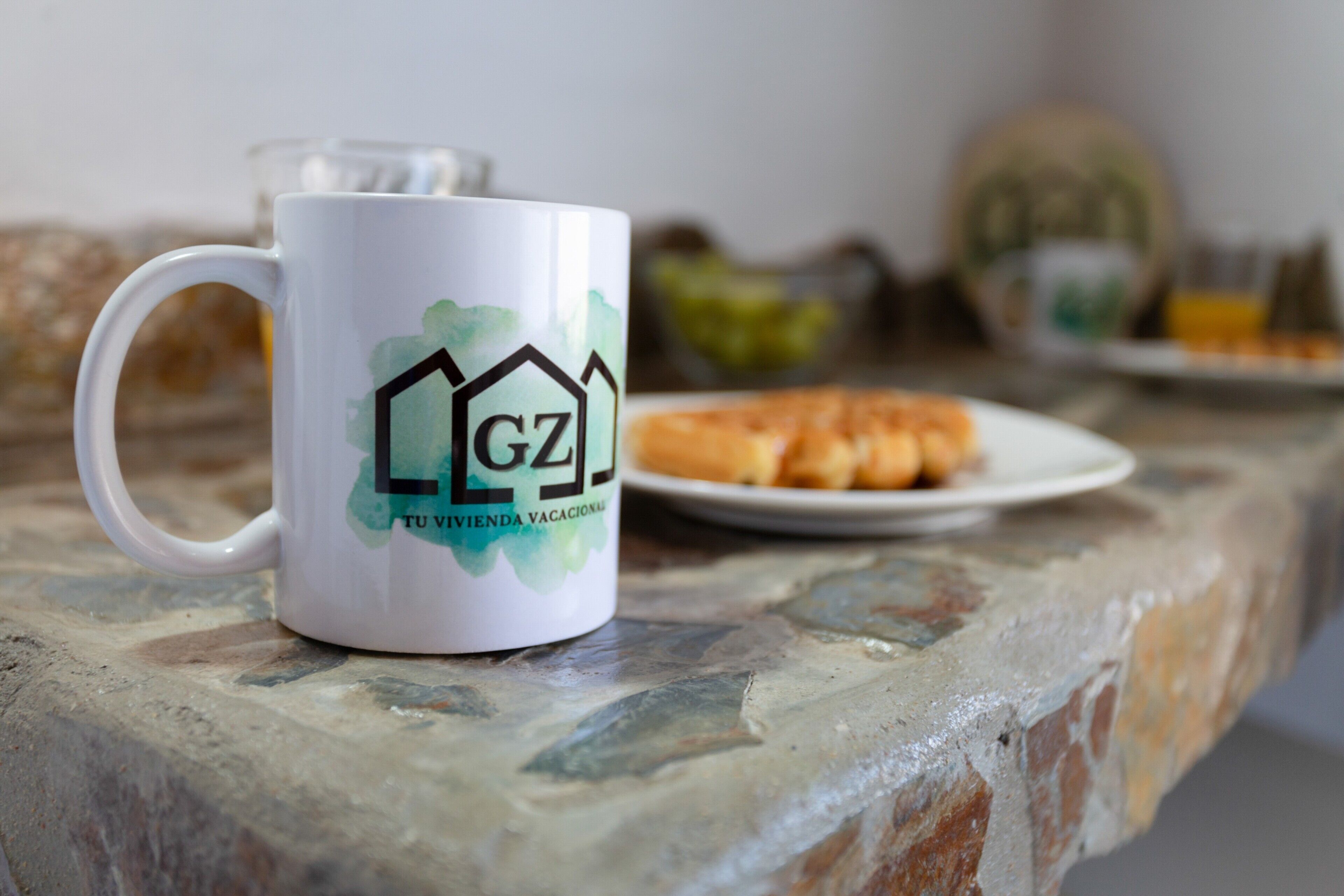 Une tasse blanche ornée d’un logo et d’un texte, posée sur une table en pierre, une assiette avec de la nourriture et un verre en arrière-plan.